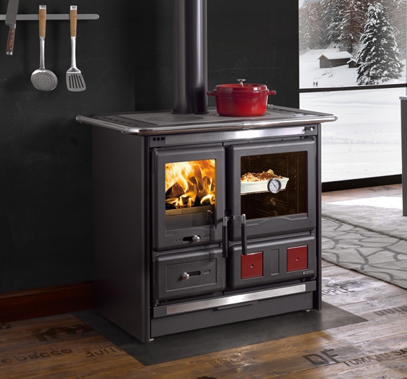 Un poêle à bois, comme le Nordica Rosa L, est doté d'une porte vitrée qui laisse entrevoir le feu chaleureux qui se dégage de l'intérieur. Un plat de cuisson repose dans un autre compartiment à droite. Des ustensiles sont suspendus au mur à gauche. Une casserole est posée sur le poêle et, à travers la fenêtre, un paysage enneigé se dévoile magnifiquement.