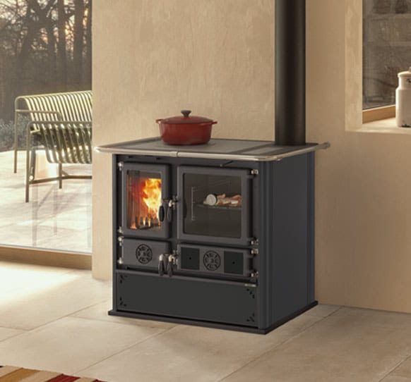 Un poêle à bois Nordica Rosa 5.0 est placé dans le coin d'une pièce avec une porte vitrée offrant une vue sur le feu. Une mijoteuse rouge repose sur les cuisinières à bois. Une fenêtre à l'arrière-plan révèle un patio extérieur avec des arbres et une chaise en métal.