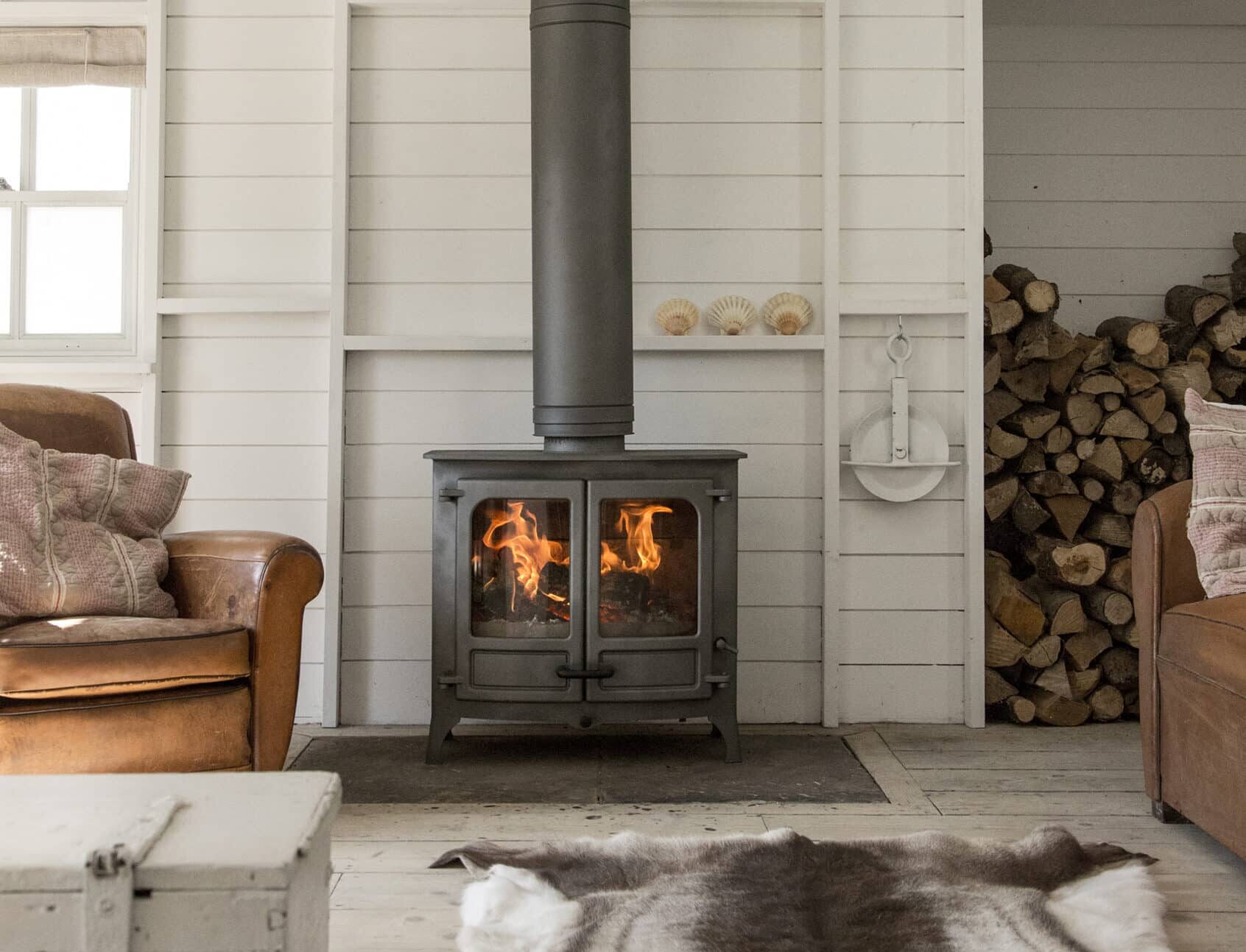 Un salon chaleureux avec un poêle à bois Charnwood Island III et un tuyau qui s'étend jusqu'au plafond. La pièce est dotée de murs en bois blanc, d'un tapis en peau de vache et de fauteuils en cuir. Le bois de chauffage est soigneusement empilé dans une alcôve à côté du poêle. Un coffre blanc fait office de table basse.