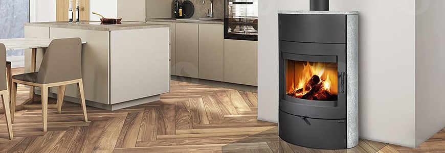 Une cuisine et une salle à manger modernes avec des meubles et des comptoirs de couleur claire. À gauche, on trouve une table à manger avec deux chaises. À droite, un poêle à bois ROMOTOP avec une façade en verre présente un feu brûlant. Le sol à motifs de bois complète parfaitement l'ambiance chaleureuse.