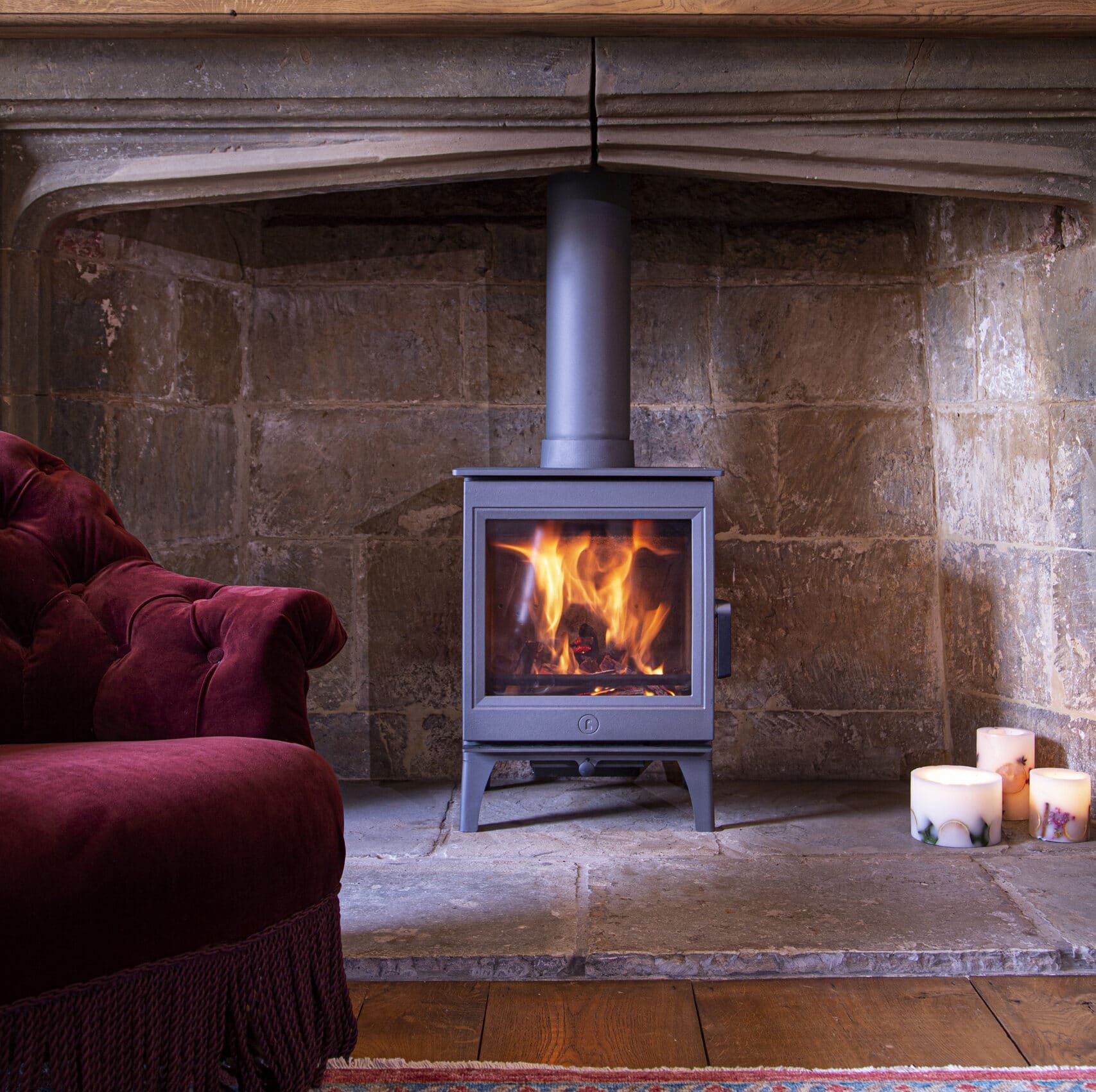 Une pièce chaleureuse est dotée d'un poêle à bois Charnwood Cranmore 5 allumé à l'intérieur d'une cheminée en pierre sous un manteau en bois. À gauche, une chaise rembourrée marron est partiellement visible. Trois bougies allumées sont placées sur le sol près de la cheminée. Un tapis à motifs recouvre le plancher en bois.