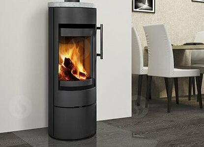 Une cuisine et une salle à manger modernes sont équipées d'un élégant poêle à bois cylindrique noir ROMOTOP avec une porte vitrée laissant apparaître un feu à l'intérieur. Le sol est en bois foncé et la salle à manger est dotée de chaises blanches et d'une table en bois. La cuisine est dotée d'armoires blanches et d'une fenêtre.